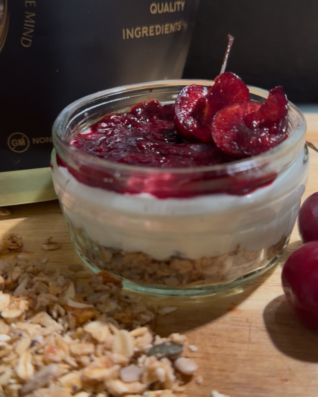 A small glass jar filled with layered Black Forest cheesecake: crunchy granola at the bottom, smooth yoghurt and cream cheese layer in the middle, and a glossy cherry compote topped with fresh cherry slices.